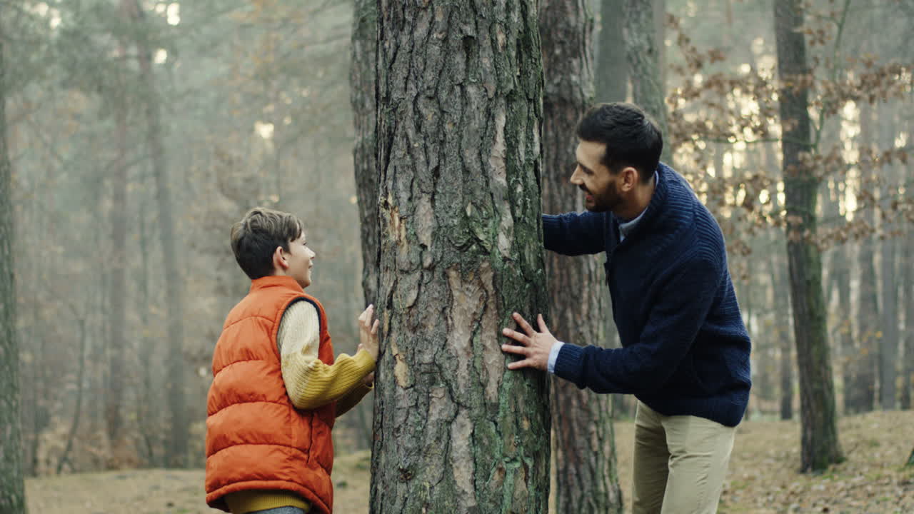 alegre padre caucásico e hijo jugando en el bosque, escondiéndose detrás de un tronco de árbol y tratando de escapar