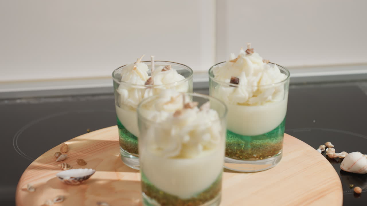Three beautifully decorated candle jars placed on round wooden tray, each layered with green and white wax and topped with whipped wax and mini shells, surrounded by scattered marine elements