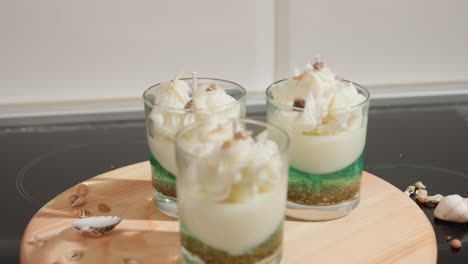 Three beautifully decorated candle jars placed on round wooden tray, each layered with green and white wax and topped with whipped wax and mini shells, surrounded by scattered marine elements