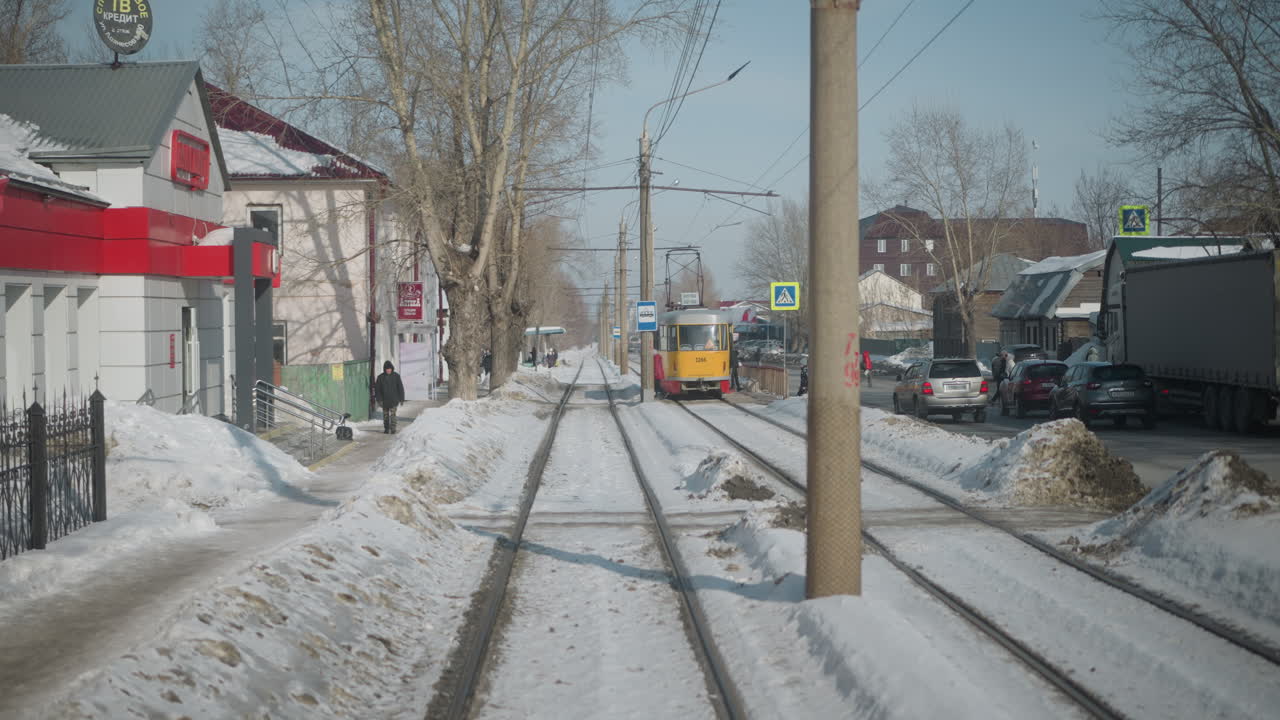 Rearward view from moving tram along snowy rails, tram stop beside road with cars, pedestrians, signage, bare trees under clear sky, urban commute scene captured in motion from carriage window