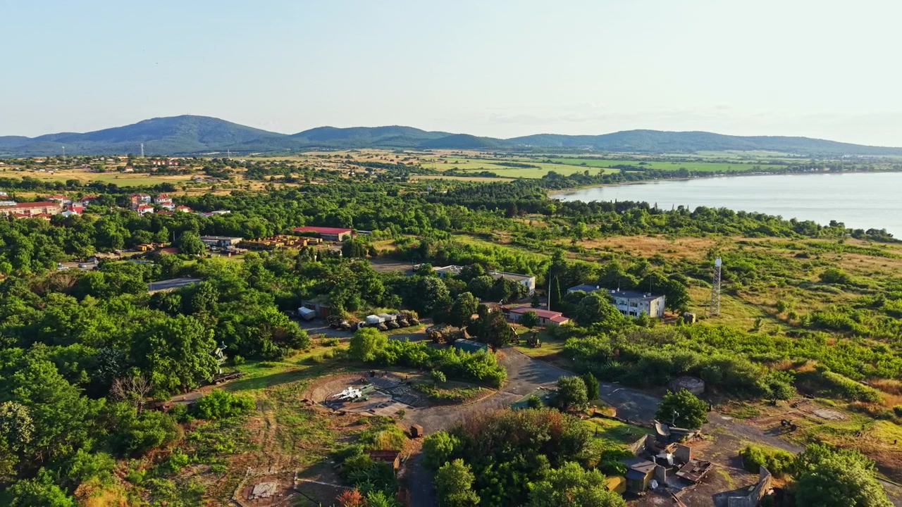 Ilmakuva vihreästä maisemasta järven lähellä, kaukaiset kukkulat taustalla, taloja ja puita aurinkoisena päivänä.