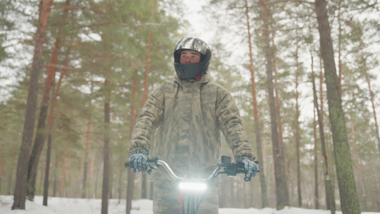 Pilota di ATV nella foresta innevata con faro luminoso che illumina la scena, giacca mimetica e guanti blu che stringono il manubrio. Sentiero ghiacciato, tracce di pneumatici nella neve, alti tronchi di pino e luce invernale attenuata creano un'atmosfera suggestiva.