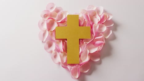 video of yellow christian cross on heart shape of pink rose petals on white background