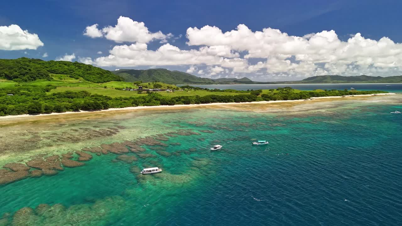 Un'immagine aerea che mostra una serena costa tropicale con campi verdeggianti, una spiaggia sabbiosa e acqua cristallina turchese piena di barriere coralline e barche in una bella giornata di sole