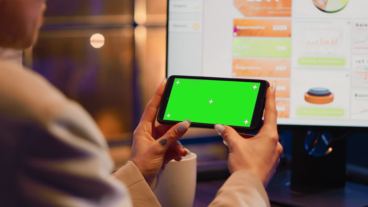 Person holding a cell phone with a green screen in front of a computer
