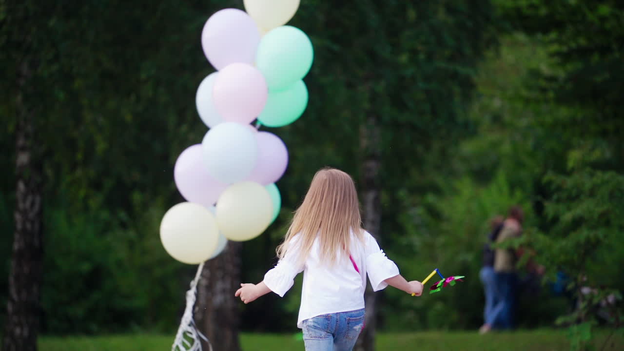 Back view of a little girl in the park. Small girl in blue jeans runs to the lots of air balloons on the natural green background in summer.