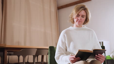 woman seated in softly lit therapy lounge reading from book with focused expression, reflecting quietly as she engages with written material in tranquil and minimal indoor setting with warm tones