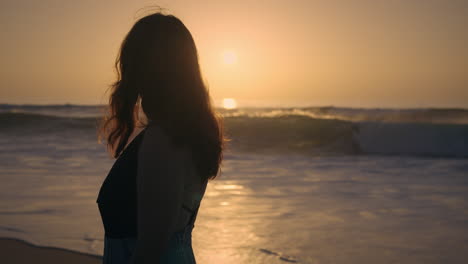 girl slowly walking by the sea at golden hour looking at the sunset while man surfs in the background medium shot