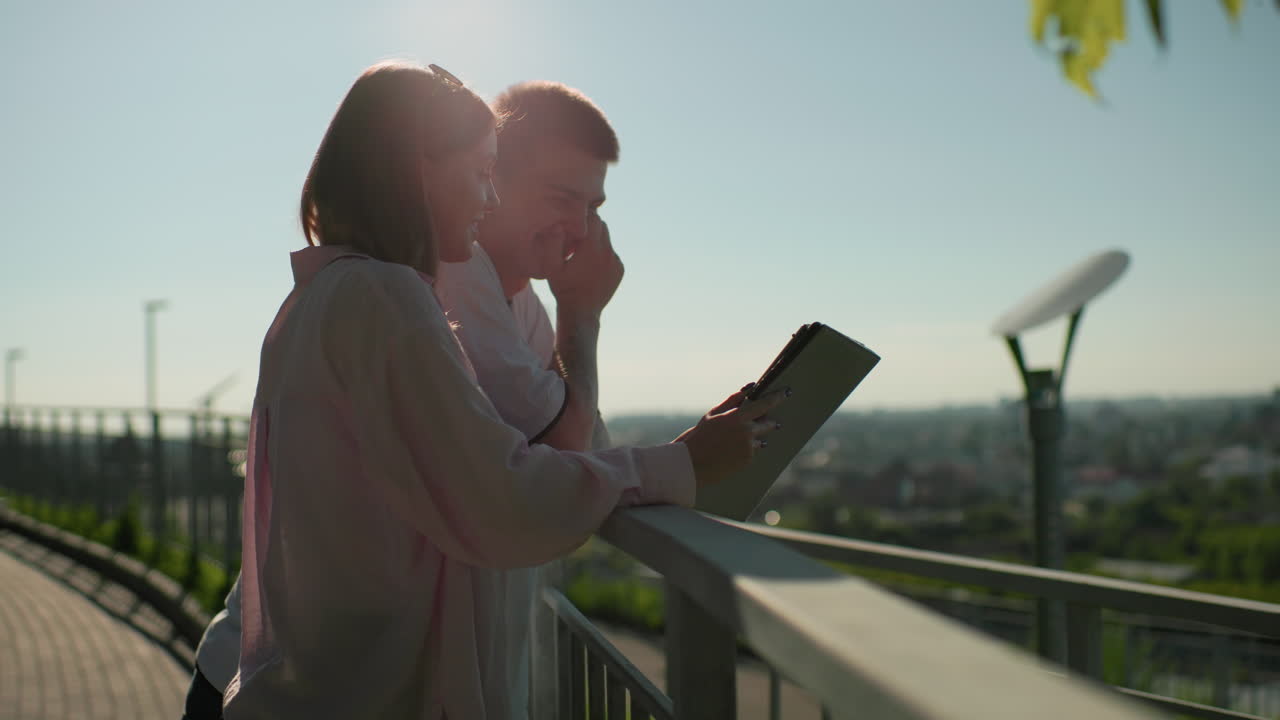 dama de camiseta rosa mostrando a su novio algo en una tableta mientras sonríen cálidamente, apoyada en un riel de hierro con una vista panorámica de la vegetación y el paisaje urbano en el fondo, bañada en la cálida luz del sol