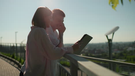 lady in pink top showing her boyfriend something on a tablet as they smile warmly, leaning on iron rail with scenic view of greenery and cityscape in background, bathed in warm sunlight