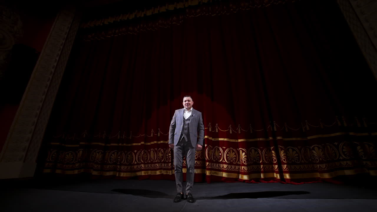 Male host talking on stage. Elegant man in grey costume performing on large scene uses gestures. Actor moving joyfully on stage.