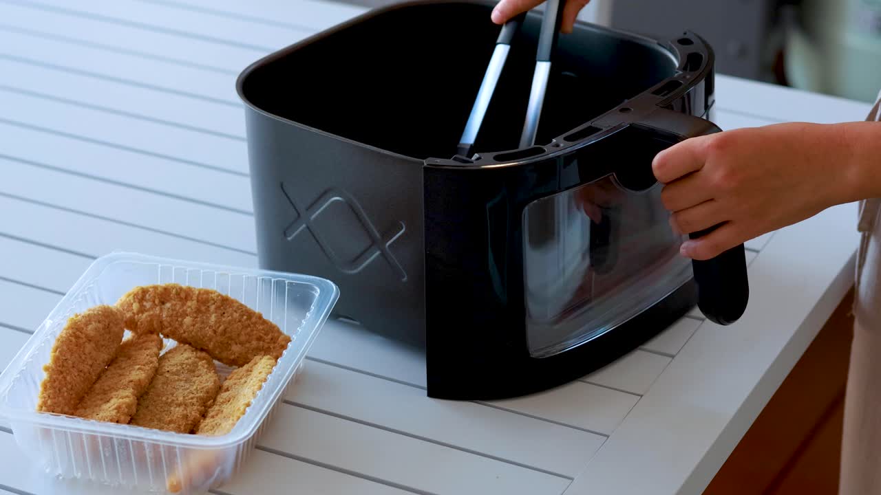 A person uses tongs to transfer breaded food pieces from a plastic tray into a black air fryer basket on a white kitchen counter under soft natural light