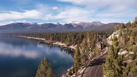 Aerial-View-of-Coastal-Road-by-Lake-Tahoe,-Docks-of-Lakefront-Homes-and-Snow-Capped-Hills