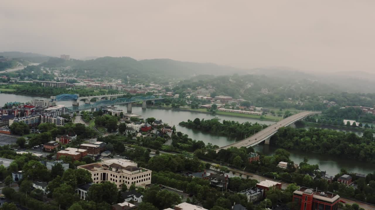 Aerial drone view of Knoxville, Tennessee featuring bridges crossing a river, surrounded by buildings, green spaces, and rolling hills. Peaceful scene capturing urban and natural beauty.