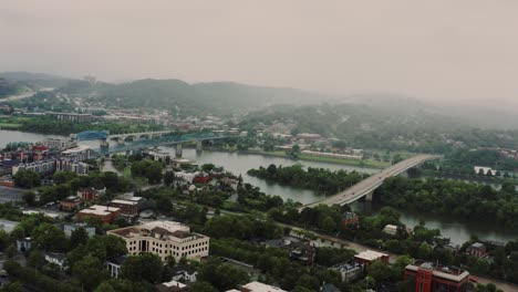 Aerial drone view of Knoxville, Tennessee featuring bridges crossing a river, surrounded by buildings, green spaces, and rolling hills. Peaceful scene capturing urban and natural beauty.