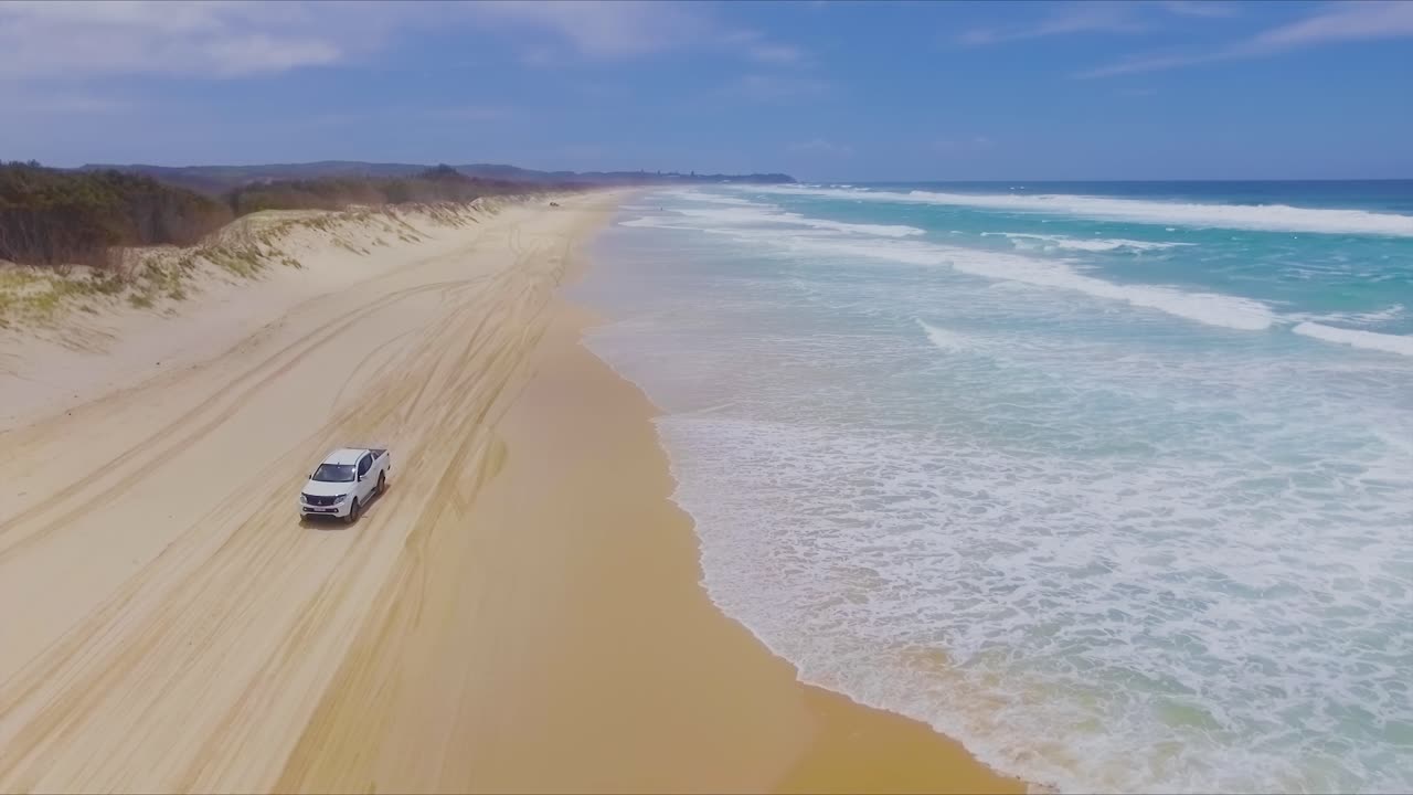 Low aerial drone shot travelling North up Main Beach on Queensland's North Stradbroke Island, as a white 4x4 four wheel drive pickup truck vehicle drives South on the beach below