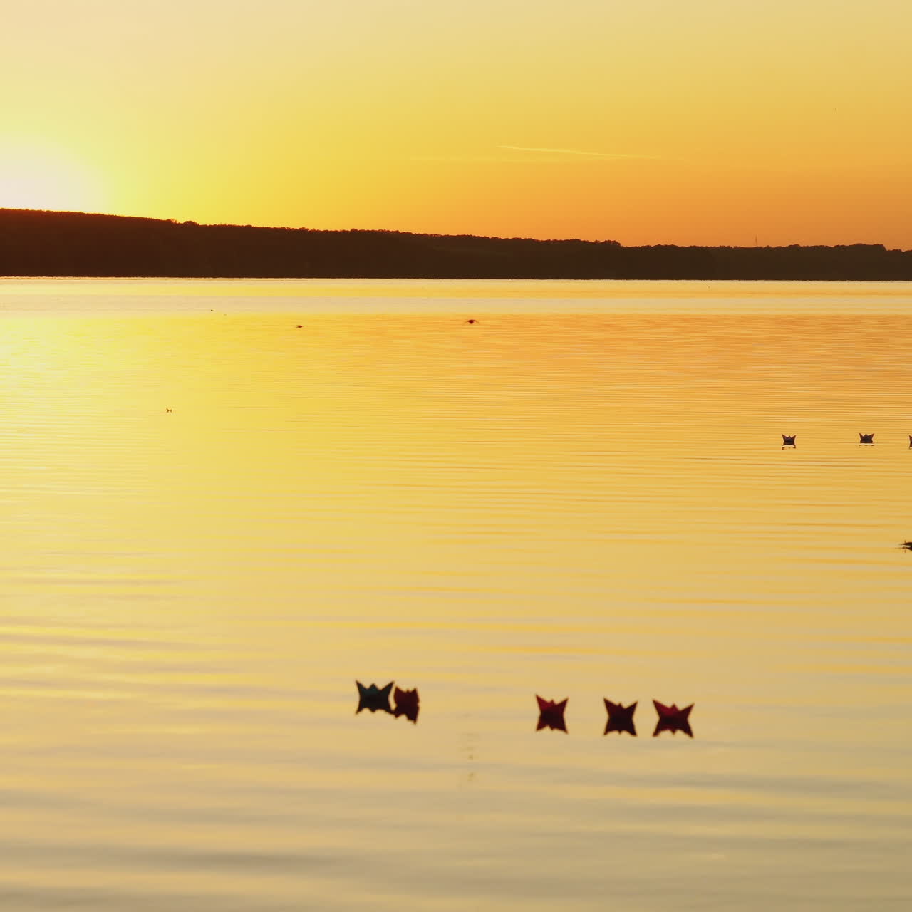 Beautiful picture of floating paper boats on the calm river at an orange sunset. Homemade origami ships on the evening water surface.