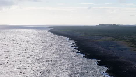 Panorámica-Aérea-De-Camiones-A-Través-De-Roca-Volcánica-Negra-Que-Se-Extiende-Hacia-Aguas-De-Mar-Abierto.