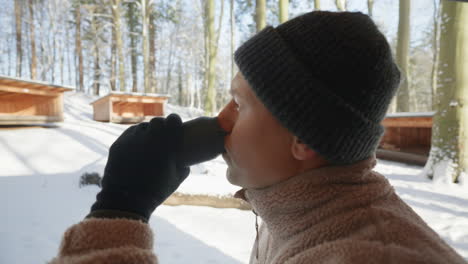 profile view of a person sipping from a thermos cup, with snowy woodland cabins in the background