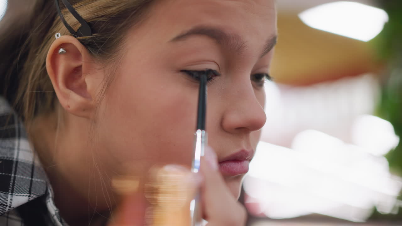 joven dibujando cuidadosamente con lápiz de ojo en su párpado con precisión, aplicación de maquillaje enfocada con una vista de cerca que destaca la atención a los detalles, capturando la rutina de belleza en un entorno casual