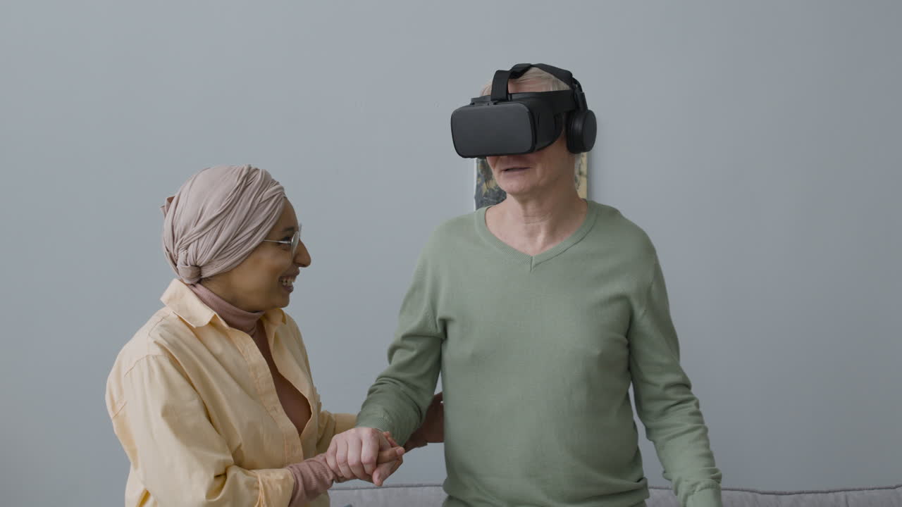 Middle Aged Arabic Woman Helping A Senior Man With Virtual Reality Glasses To Stand Up From The Couch At Home