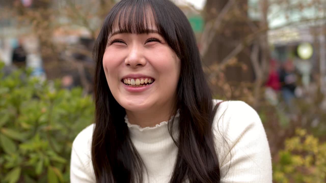 하얀 스웨터를 입은 쾌활한 아시아 여성이 야외 인터뷰 중에 웃고 미소 짓는다