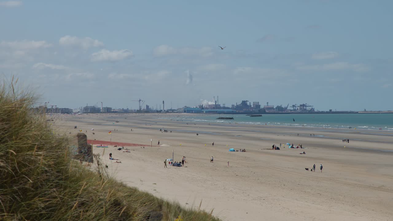 Wide shot of sandy beach, people relaxing, distant industrial port, natural light, slight camera pan