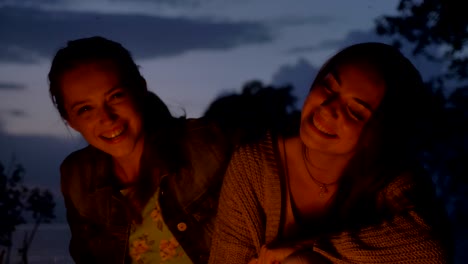 two young caucasian girls sit in the evening by the fire in the nature, laughing, smiling