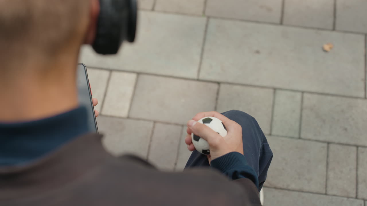 Uomo bianco ripreso dall'alto che controlla il telefono mentre tiene in mano un mini pallone da calcio su una panchina sul marciapiede, pollice che scorre su un'app per seguire i risultati in diretta, vibrazione di notifica, energia da spettatore urbano, abbigliamento casual