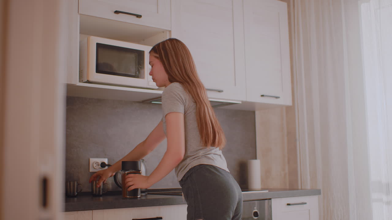 Una donna che prepara la colazione in cucina apre il pensile per prendere un contenitore di bevande, lo appoggia sul bancone, sposta una tazza in avanti, apre il contenitore e prepara la bevanda con un cucchiaio, il tutto illuminato dalla luce del giorno.