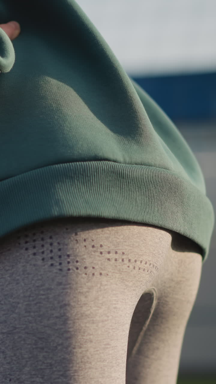 primer plano de una joven con sudadera verde y cabello largo realizando ejercicios de torsión de la cintura, fondo borroso con red deportiva y edificio en una cancha de fitness al aire libre