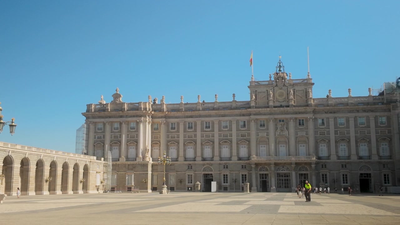 Una vista ampia dello storico Palazzo Reale di Madrid e dei grandi archi della Piazza d'Armi sotto un cielo azzurro e limpido. Perfetto per documentari di viaggio e contenuti sul turismo europeo.