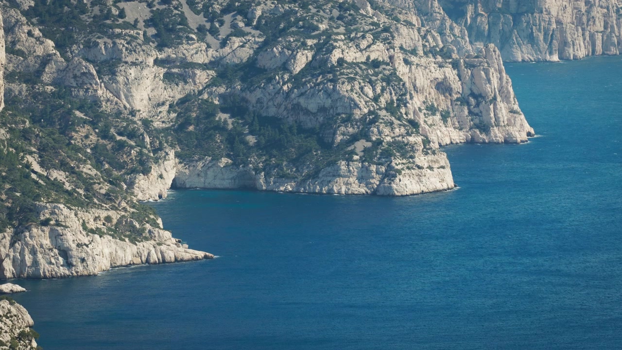 Il paesaggio aspro del Parco Nazionale delle Calanques in Francia. L'acqua di un blu profondo si infrange contro le scogliere bianche, che sono coperte da alberi e arbusti. Il sole è basso nel cielo