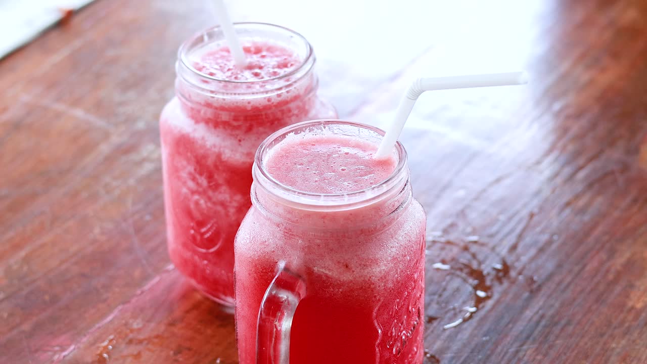 Hands reach for vibrant watermelon juice in glass mugs on a wooden table. Bright lighting enhances the refreshing atmosphere