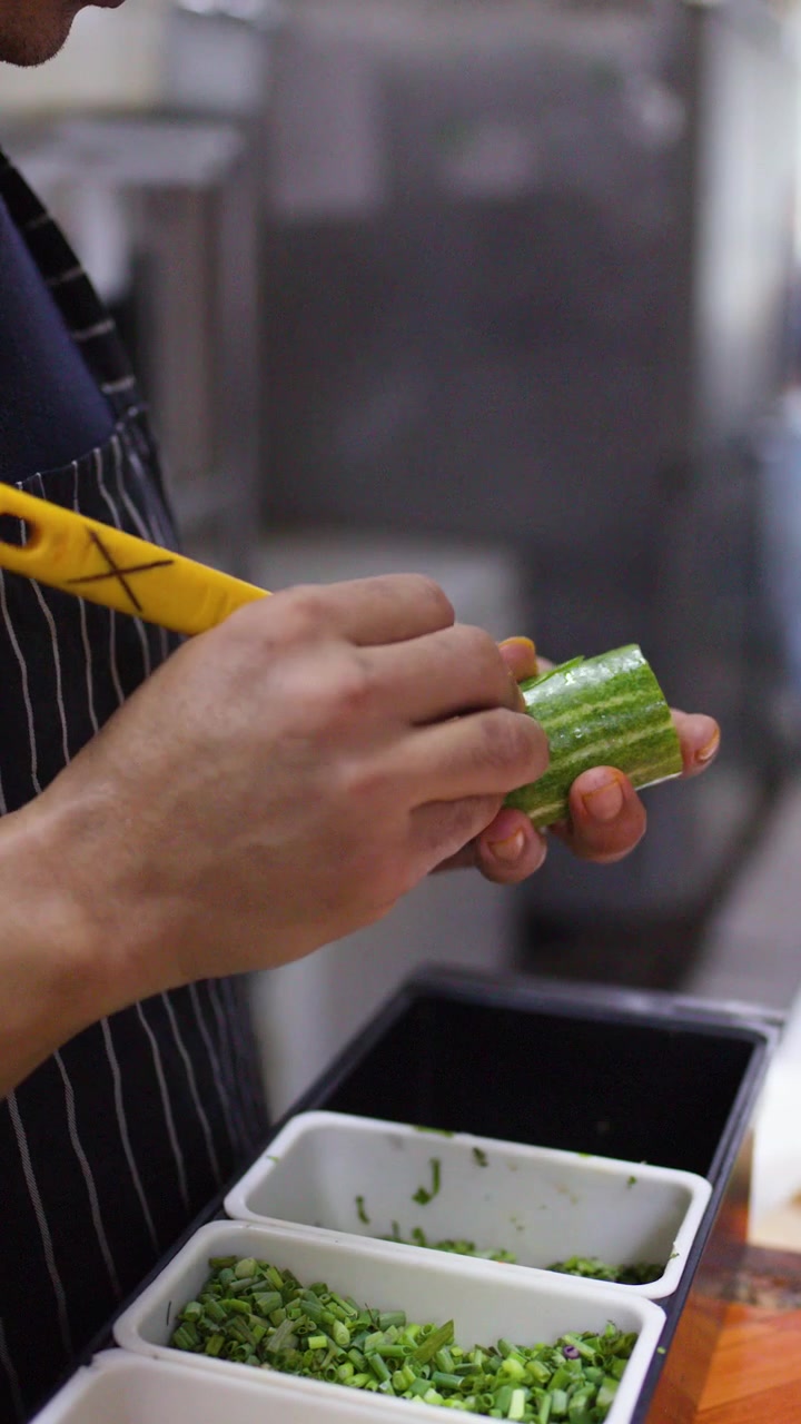 Chef professionista con grembiule a righe usa un coltello affilato per affettare un cetriolo sopra contenitori per alimenti