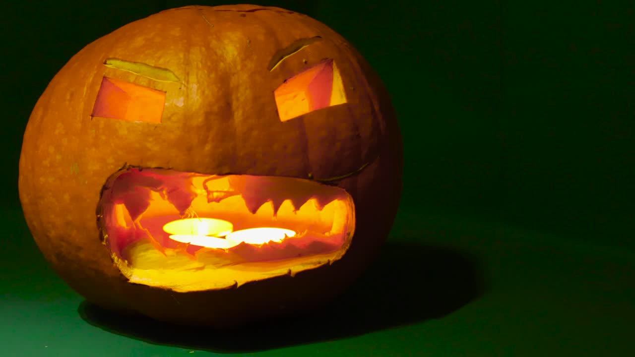 A carved out Halloween pumpkin on a green studio background or a green screen while the camera is moving around it. Candles are lit and glowing inside of the pumpkin through its scary face. decoration