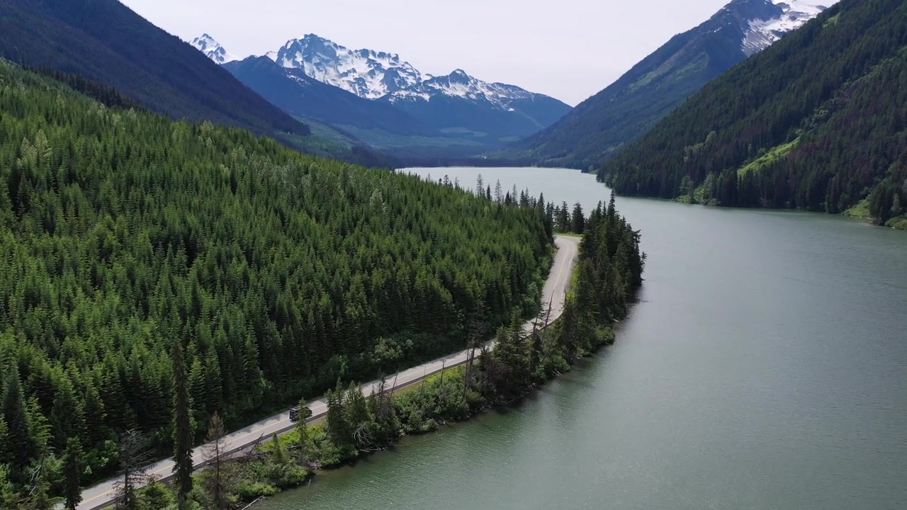 Ripresa aerea con drone dall'alto di un furgone che viaggia su una strada asfaltata attraverso dolci colline e campi verdi. Una scena cinematografica di un viaggio on the road in stile "van life" nell'entroterra della Columbia Britannica sotto un sole splendente nel pomeriggio.