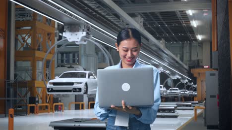 trabajadora profesional asiática de pie con su portátil en una fábrica de automóviles ev, instalación industrial de alta tecnología, trabajo continuo
