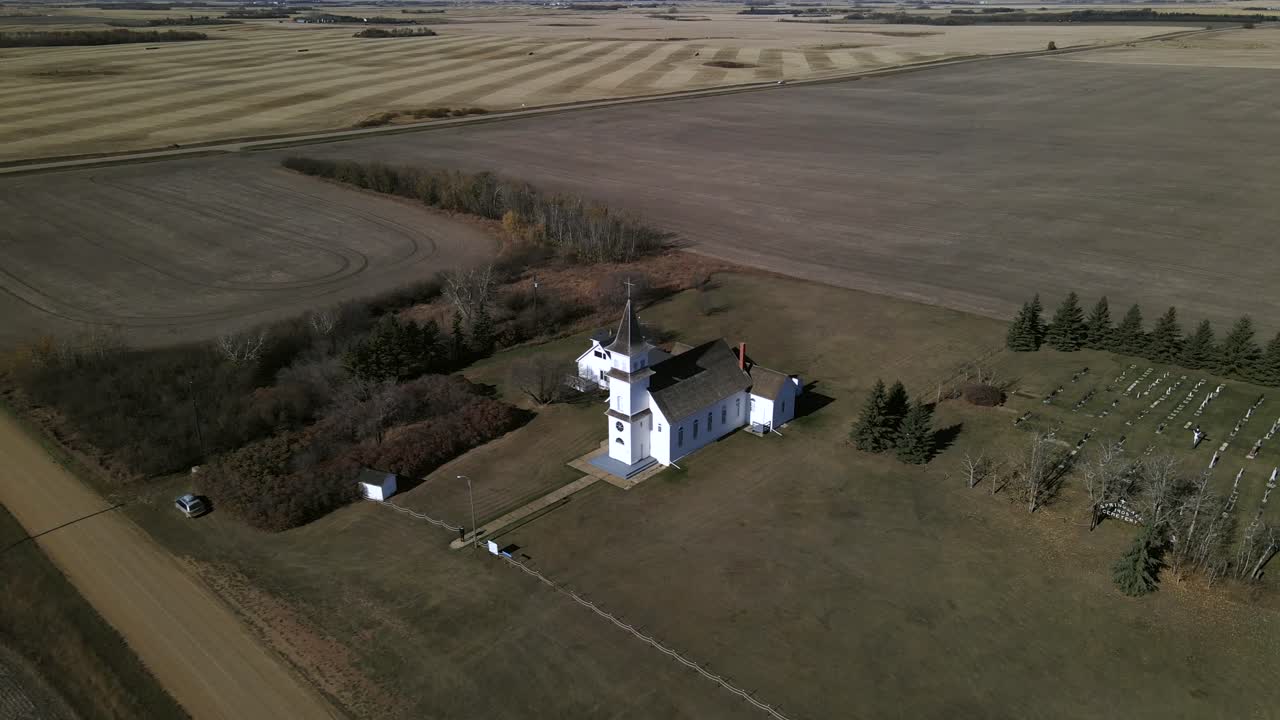 imágenes de drones de la hermosa y antigua iglesia patrimonial sentada aislada en el paisaje de las praderas de américa del norte mientras un solo automóvil conduce a lo largo de una carretera recta en el fondo