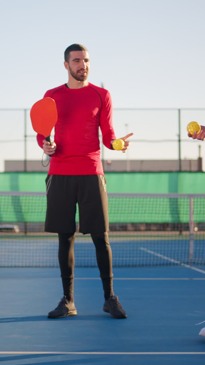 A man in a red shirt teaching two women with curly hair how to play pickleball on a blue court on a sunny day. Vertical