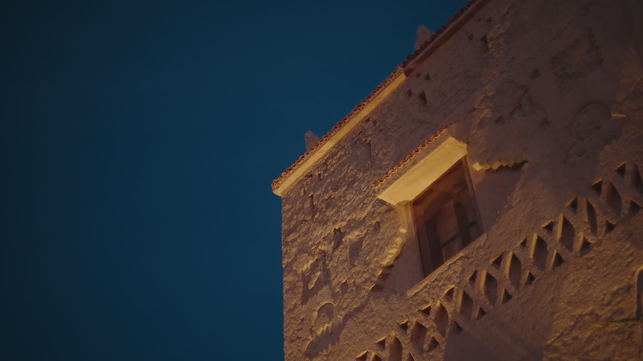 Vista dal basso di una torre kasbah in argilla illuminata con decorazioni geometriche intagliate e davanzale in legno, contro un cielo notturno blu intenso.