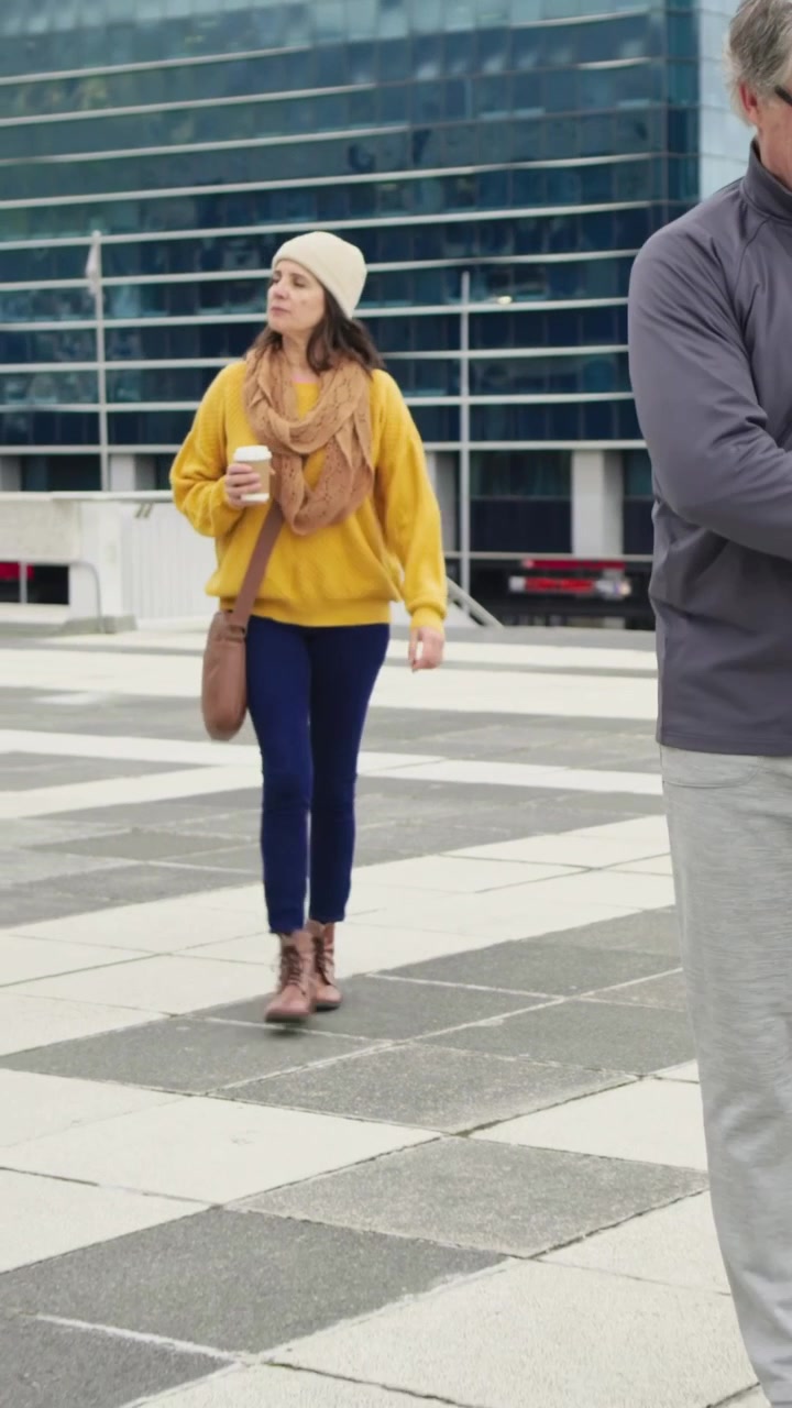 Vertical video: Entering frame, man adjusting bag crossing plaza, woman in yellow holding coffee