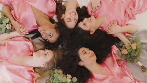 top view of group of multiethnic female friends and bride dressed in pink and white silk nightdresses holding hands with bouquets and laughing while laying on floor