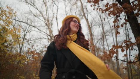 stylish woman in yellow beret and sunglasses playfully touches muffler while strolling through autumn forest, enjoying the crisp air and colorful leaves in a serene outdoor setting