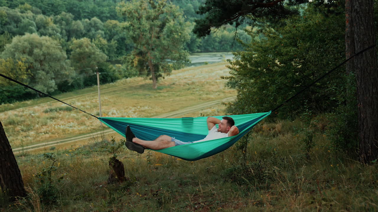 Happy Caucasian man lies comfortably in a hammock hands behind his head. Relaxed person looks dreamily up swaying slowly in hammock. Nature at backdrop.