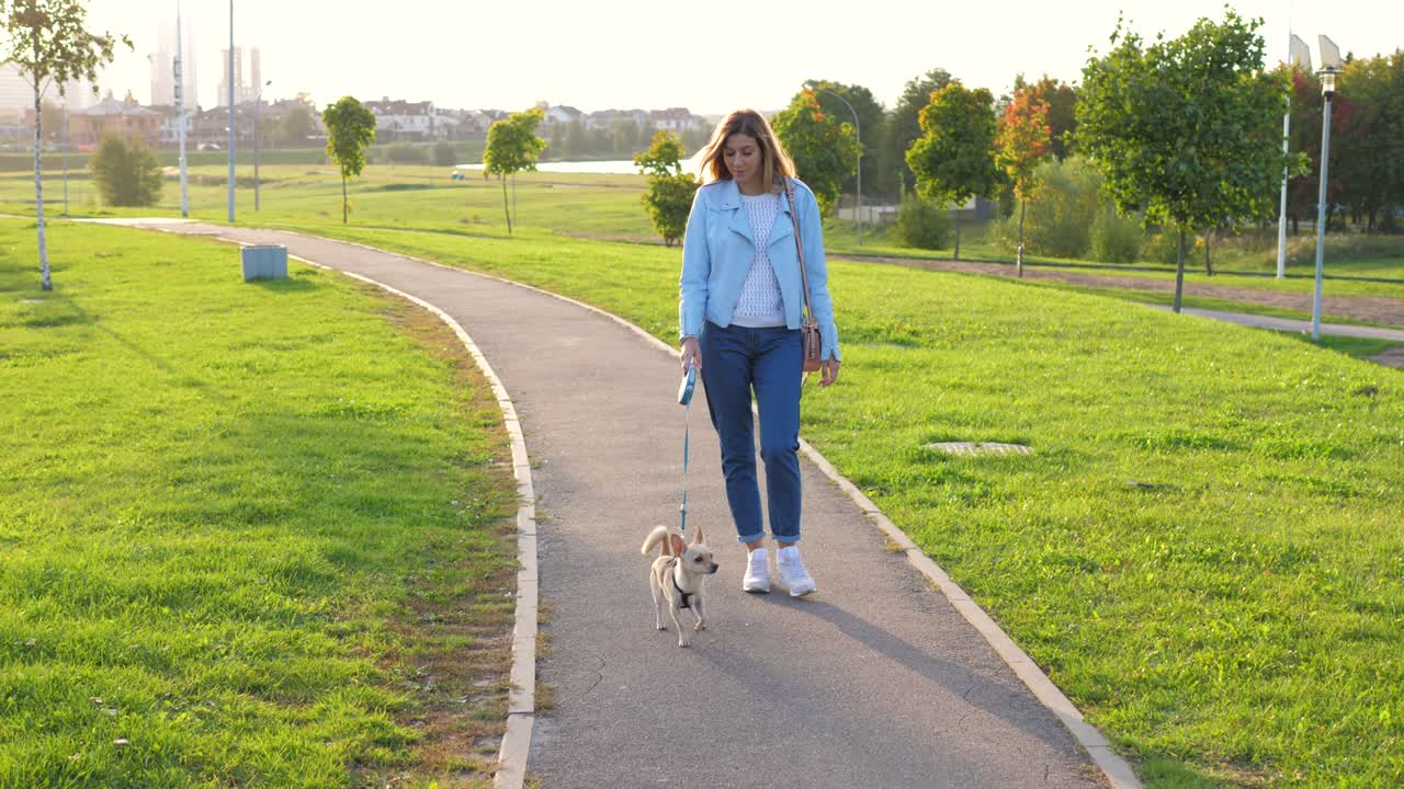 mujer caucásica camina afuera con su pequeño perro al atardecer en un parque verde