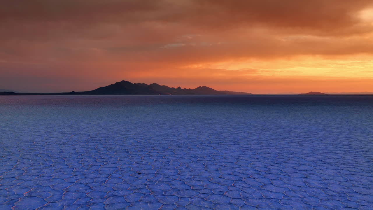 Footage over the salty landscape colored blue at sunset. Approaching black silhouette of mountains against orange sky. Bonneville Salt Flats, Utah, United States