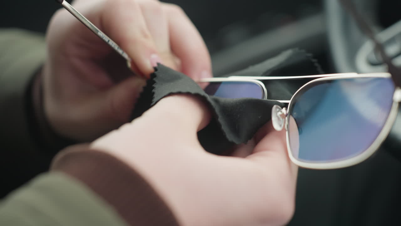 Close up of person cleaning glasses lens with black cloth inside car cabin showing hand wiping reflective optics near steering wheel under natural light capturing maintenance motion