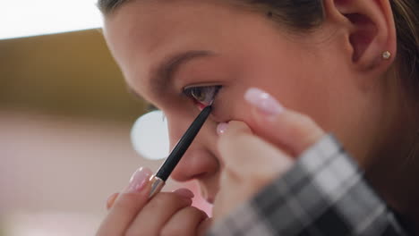 close-up view of young lady gently applying eye pencil on eyelid while using her hand to bring down her eye for precise makeup application, focusing on delicate eye makeup process