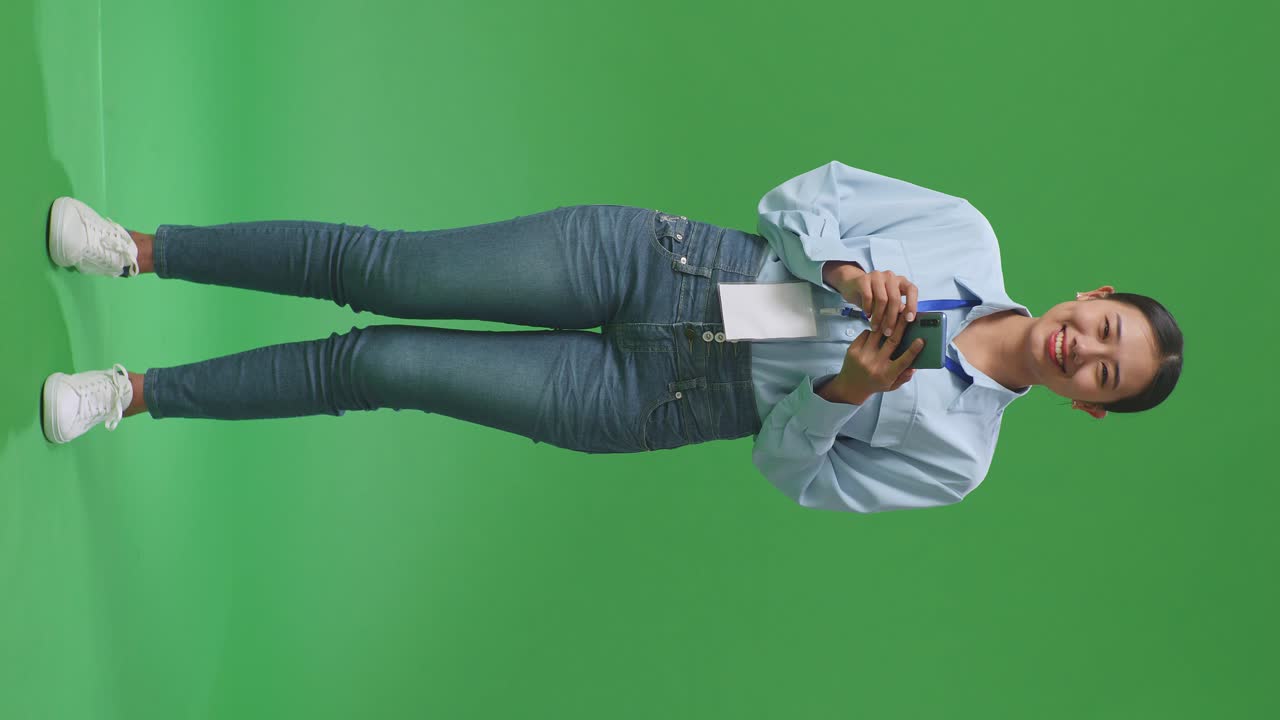 cuerpo lleno de una mujer de negocios asiática usando un teléfono móvil y sonriendo a la cámara en el fondo de la pantalla verde en el estudio